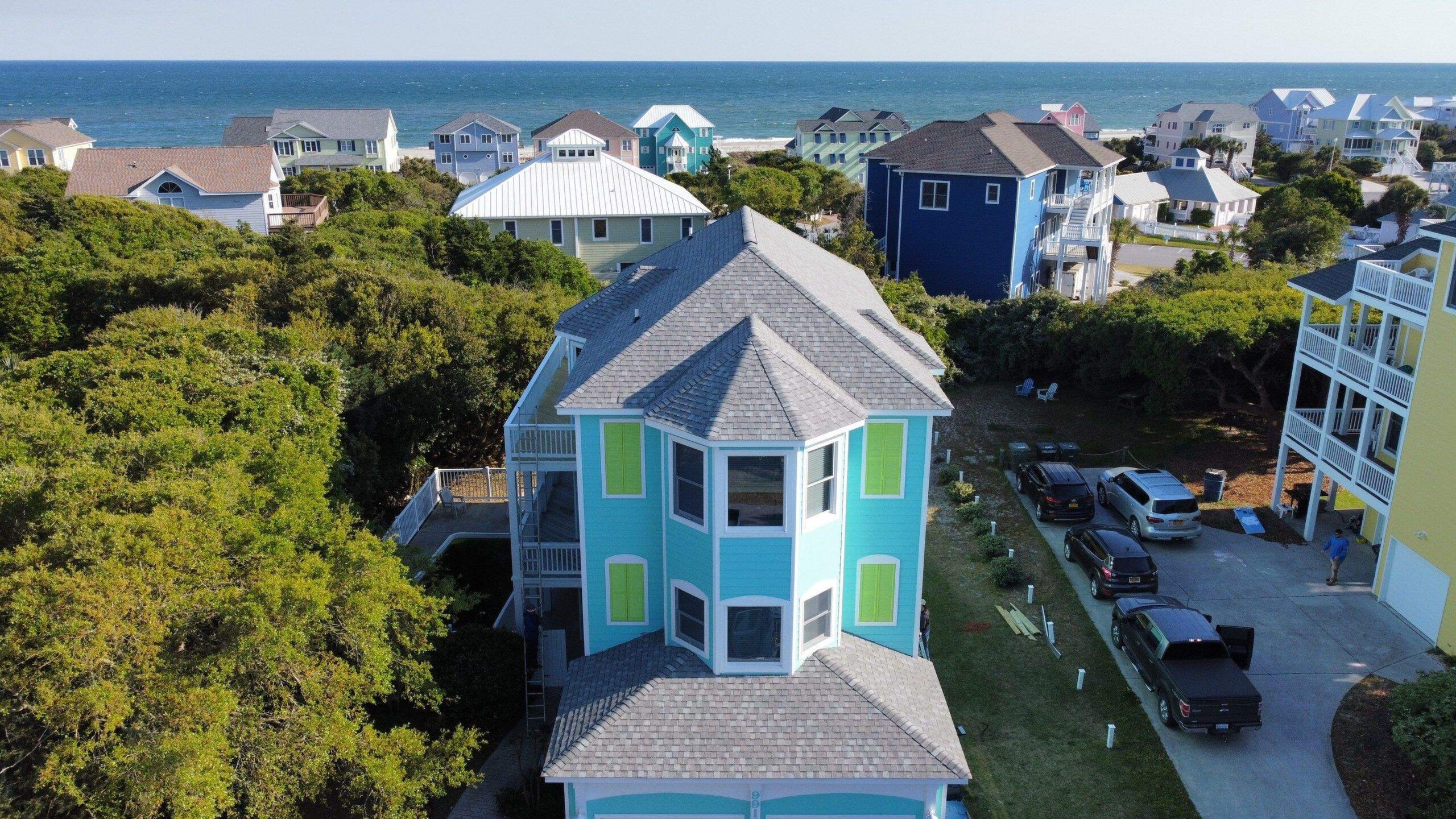 A new shingle roof on a bright blue beach house, with the ocean and other beach houses behind it