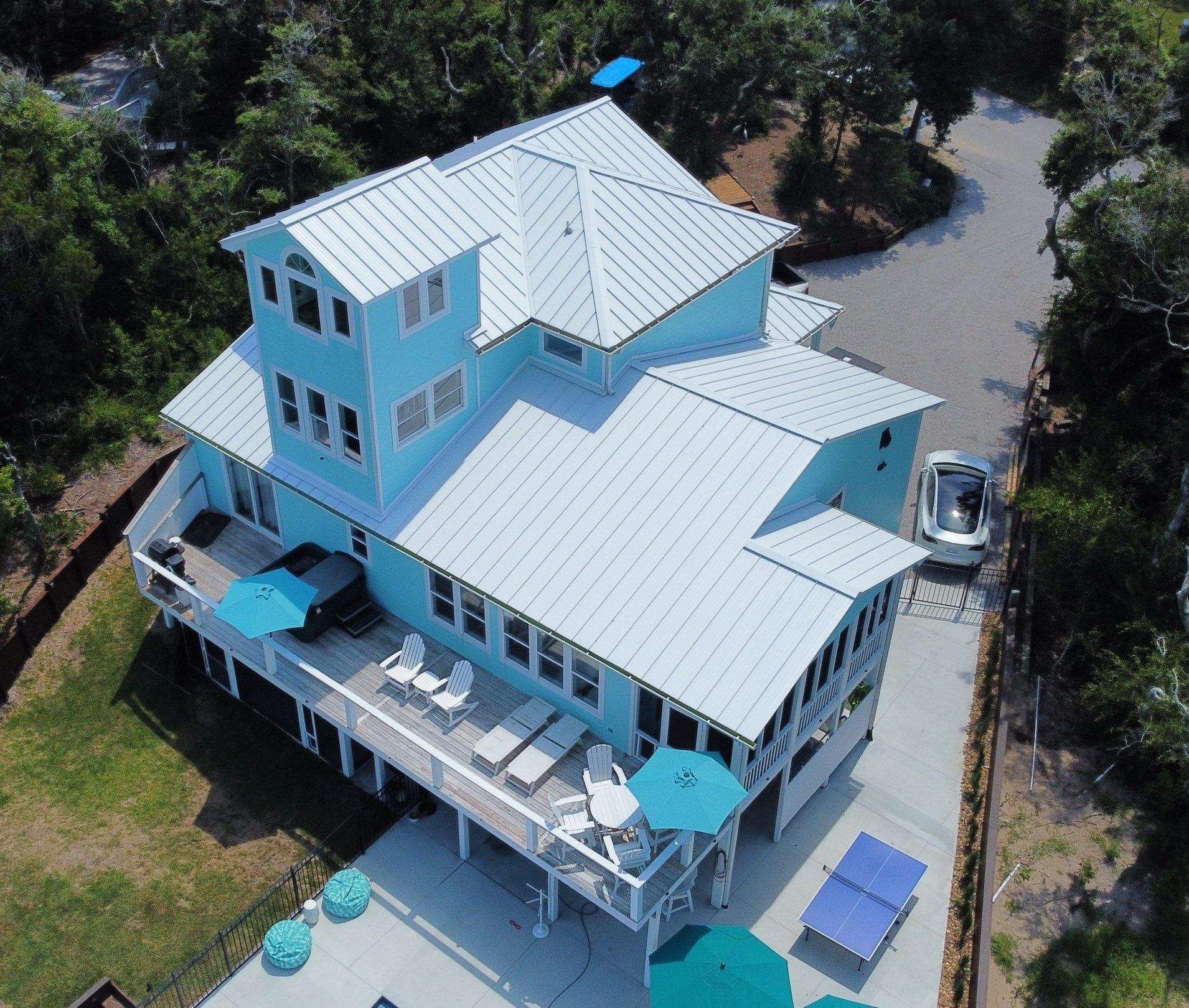 A turquoise coastal house with a new standing seam metal roof and a wide deck, seen from a drone